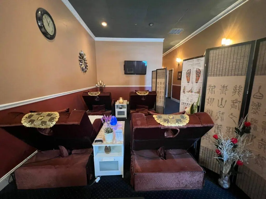 Cozy foot massage area at Hong's Massage featuring two comfortable reclining chairs with foot basins, separated by a small table. Warm lighting, decorative wall clock, and traditional Chinese medicinal charts create a relaxing atmosphere."