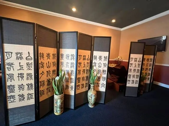 "Reception area of Hong's Massage featuring elegant room dividers with traditional Chinese calligraphy. Warm lighting illuminates beige walls and dark blue carpeting. Decorative plants in vases add a touch of nature to the serene atmosphere."